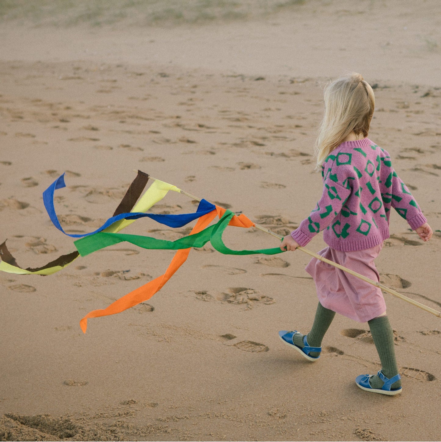 Child walking on beach holding colourful flag wearing pink and green wool cardigan.